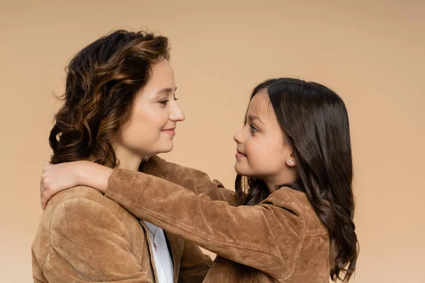 Side view of mother and daughter in suede jackets smiling at each other isolated on beige — Stock Photo