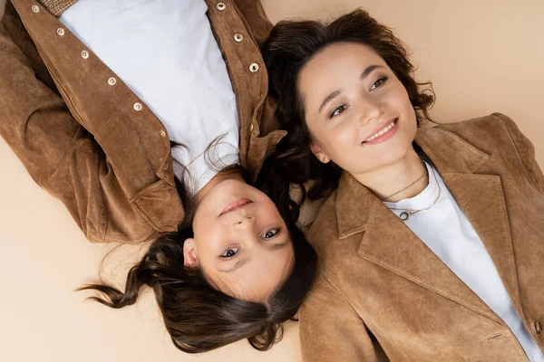 Top view of brunette girl lying near mom in trendy autumn jacket and smiling at camera on beige background — Stock Photo