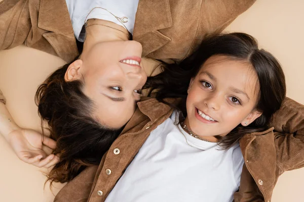 Draufsicht einer glücklichen und stilvollen Frau, die ihre Tochter lächelnd in die Kamera sieht und auf beigem Hintergrund liegt — Stockfoto