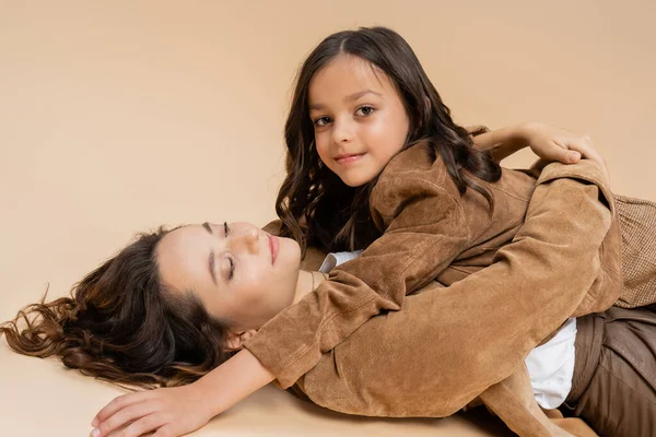 Stylisches Mädchen lächelt in die Kamera neben Mama, die mit geschlossenen Augen auf beigem Hintergrund liegt — Stockfoto