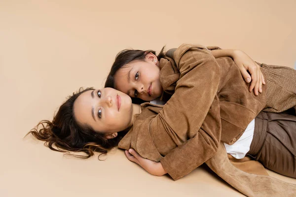 Mamá positiva y la hija en chaquetas de otoño de moda tumbado y mirando a la cámara en fondo beige - foto de stock