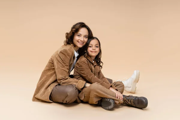 Mujer feliz y chica en botas de moda y chaquetas de gamuza marrón sentado sobre fondo beige - foto de stock