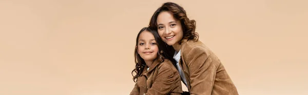 Mujer alegre en chaqueta de otoño mirando a la cámara cerca de la hija de moda aislado en beige, pancarta - foto de stock