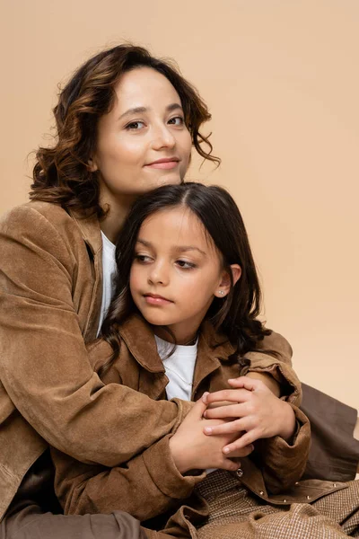 Felice ed elegante donna abbracciando figlia in camoscio marrone giacca isolata su beige — Foto stock