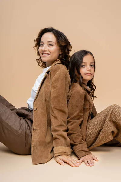 Mujer alegre en traje de otoño de moda sentado espalda con espalda con hija y mirando a la cámara en fondo beige - foto de stock