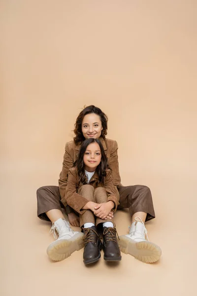Smiling mom and daughter in brown suede jackets and boots posing on beige background — Stock Photo