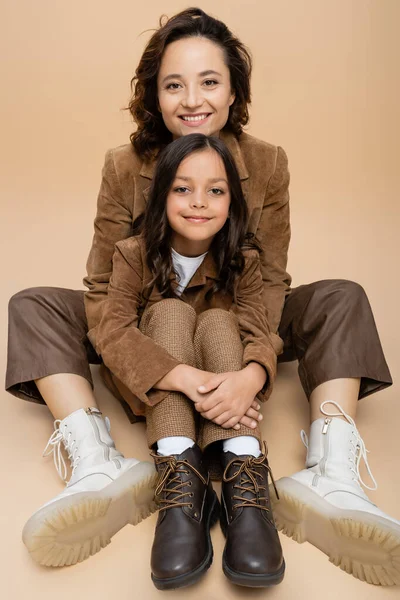 Felice donna e bambino in camoscio giacche e stivali alla moda seduto su sfondo beige — Foto stock