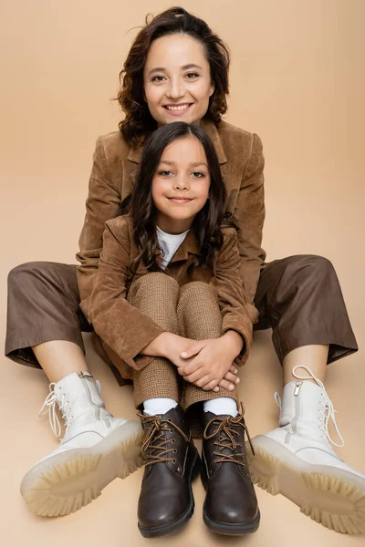 Mujer feliz y niño en chaquetas de gamuza y botas de moda sentado sobre fondo beige - foto de stock