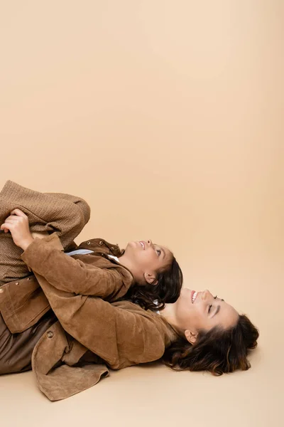 Vista lateral da menina se divertindo e deitado na mãe feliz em roupas de outono no fundo bege — Fotografia de Stock