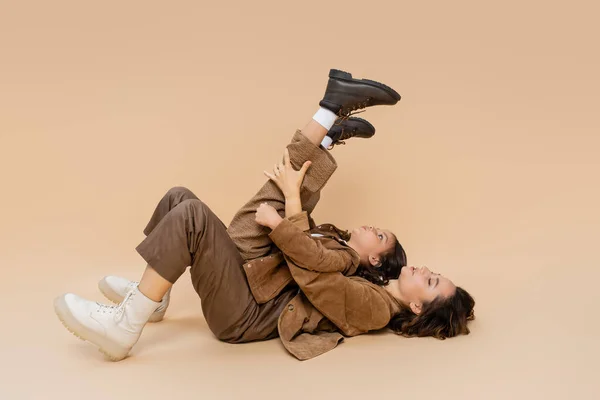 Side view of girl with raised legs having fun and lying on mom in autumn outfit on beige background — Stock Photo