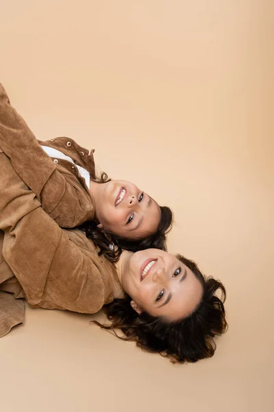 Top view of cheerful woman and child in brown suede jackets lying and looking at camera on beige background — Stock Photo