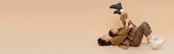 Vista laterale della ragazza in stivali alla moda sdraiato su mamma con gambe sollevate mentre si diverte su sfondo beige, banner — Foto stock