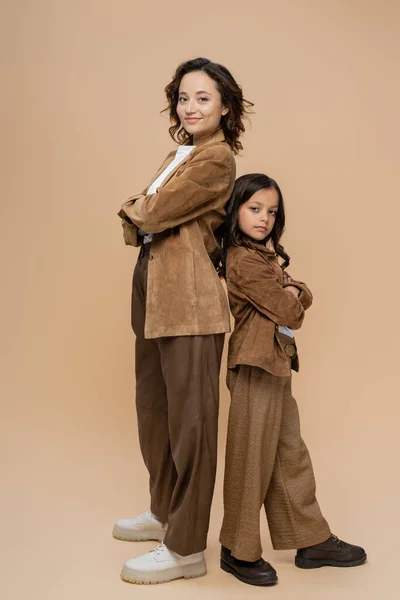 Full length of fashionable mother and child standing back to back with crossed arms on beige background — Stock Photo