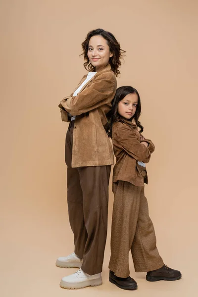 Toute la longueur de la mère à la mode et l'enfant debout dos à dos avec les bras croisés sur fond beige — Photo de stock