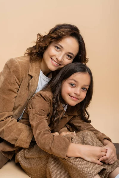 Pleased mother and daughter in brown suede jackets looking at camera on beige background — Stock Photo