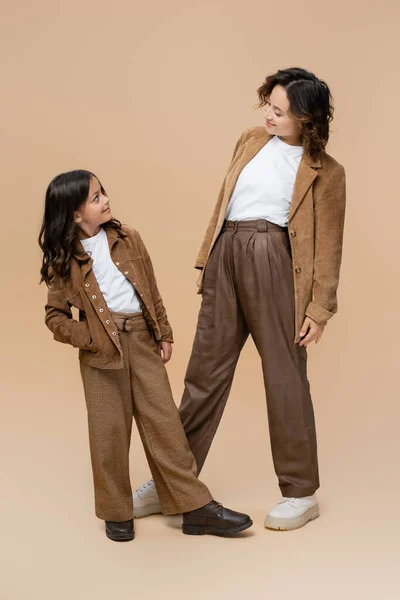 Cheerful mom and daughter in suede jackets and trendy pants looking at each other on beige background — Stock Photo