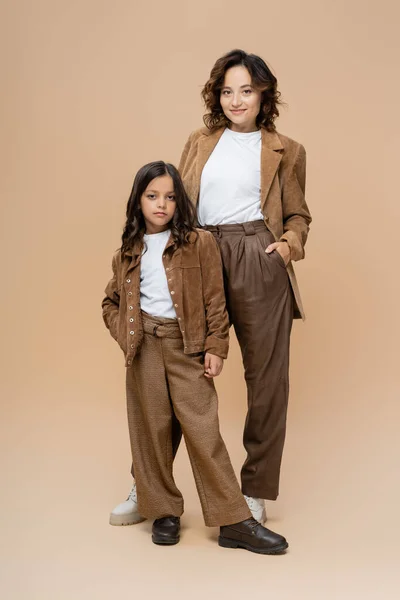 Longitud completa de la mujer y la niña en ropa de otoño de moda posando con las manos en los bolsillos sobre fondo beige - foto de stock