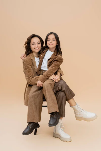 Happy mother and daughter in trendy autumn outfit and boots sitting on chair on beige — Stock Photo