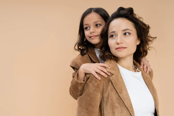 Brunette girl hugging shoulders of mom in brown suede jacket while looking away isolated on beige — Stock Photo