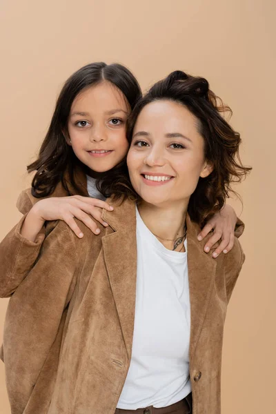 Brunette girl hugging shoulders of happy mom in brown suede jacket isolated on beige — Stock Photo