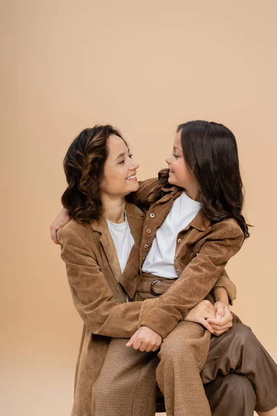Joyful mom and daughter in trendy autumn outfit looking at each other isolated on beige — Stock Photo
