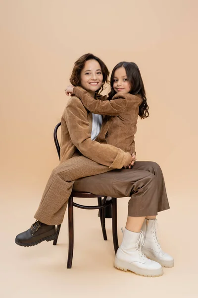 Donna e bambino alla moda in abito autunnale abbracciati sulla sedia e sorridenti alla macchina fotografica su sfondo beige — Foto stock