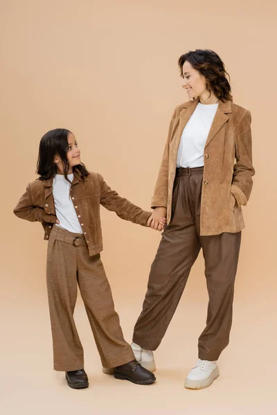 Full length of woman and girl in trendy autumn outfit holding hands and smiling at each other on beige background — Stock Photo