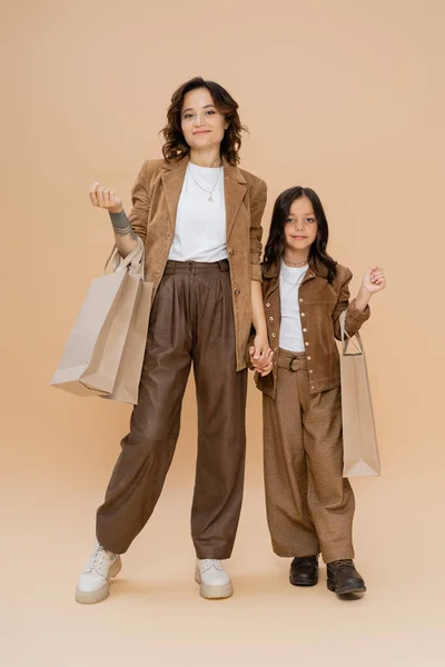 Full length of woman and girl in stylish autumn clothes standing with shopping bags on beige background — Stock Photo