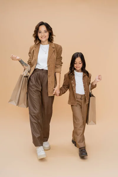 Mère et enfant dans des vêtements d'automne à la mode tenant la main tout en marchant avec des sacs à provisions sur beige — Photo de stock