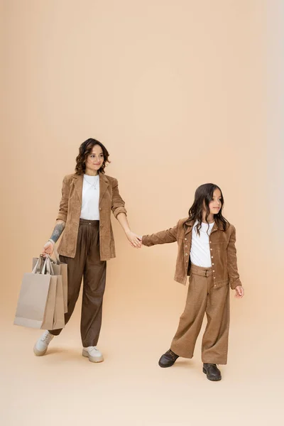 Full length of girl in trendy clothes holding hands with happy mom with shopping bags on beige background — Stock Photo