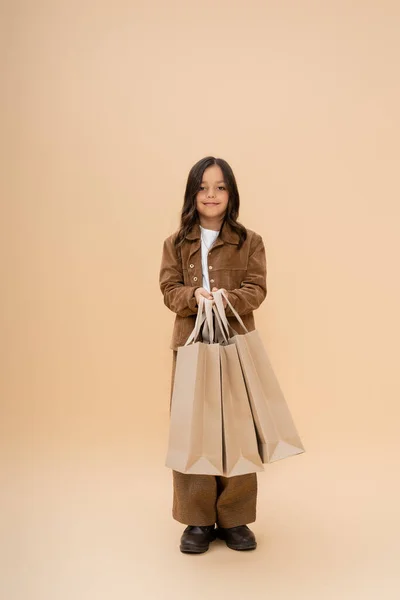 Menina sorridente na moda roupas de outono segurando sacos de compras e olhando para a câmera no fundo bege — Fotografia de Stock