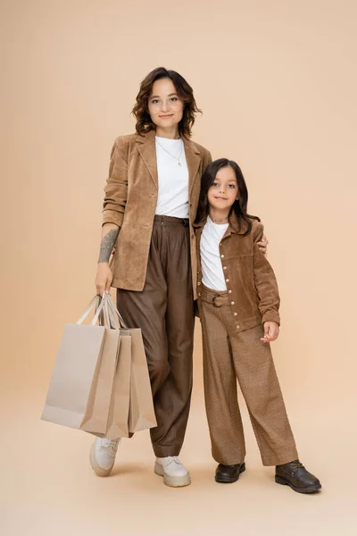 Frau in voller Länge mit Einkaufstaschen umarmt Tochter, während sie im trendigen Herbst-Outfit auf beige posiert — Stockfoto