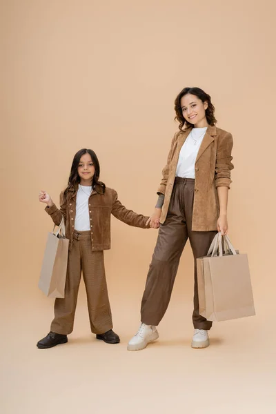 Happy mom and daughter in trendy autumn outfit holding hands and shopping bags on beige — Stock Photo