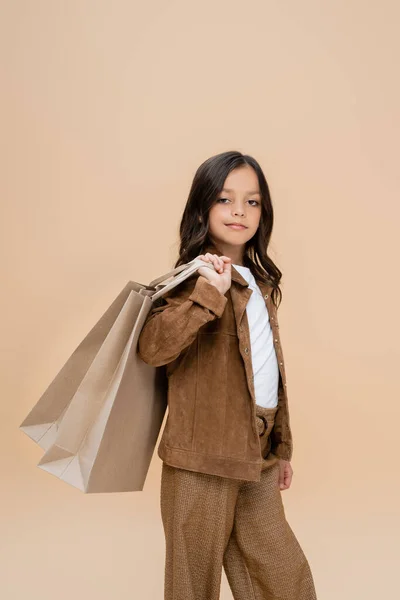 Bambino in giacca scamosciata e pantaloni marroni in posa con shopping bags isolati su beige — Foto stock