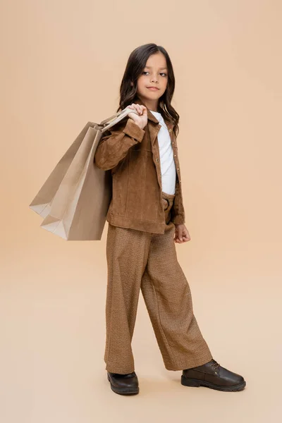 Pleine longueur de fille en veste en daim et pantalon marron tenant des sacs à provisions sur fond beige — Photo de stock