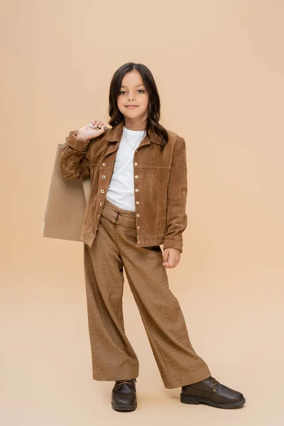 Brunette girl in suede jacket and brown pants posing with shopping bags on beige background — Stock Photo