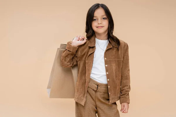 Morena chica con bolsas de compras posando en chaqueta de gamuza y mirando a la cámara aislada en beige - foto de stock