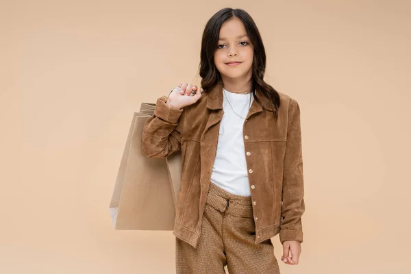 Brünettes Mädchen mit Einkaufstaschen posiert in Wildlederjacke und blickt vereinzelt in die Kamera auf beige — Stockfoto