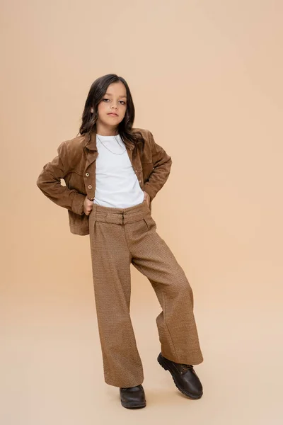 Full length of trendy girl in brown pants and suede jacket standing with hands on hips on beige background — Stock Photo