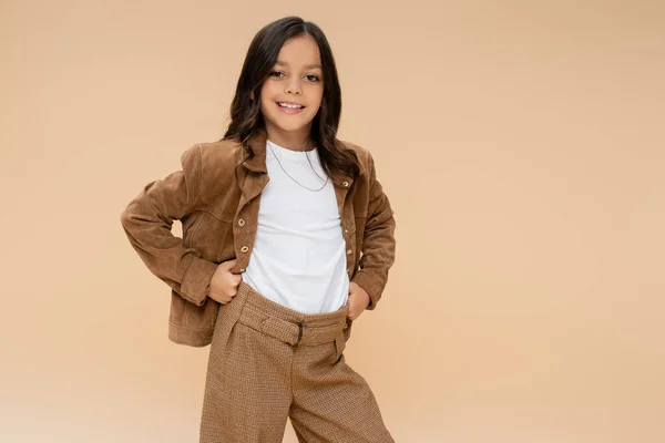 Niño alegre en traje de otoño de moda de pie con las manos en las caderas aisladas en beige - foto de stock