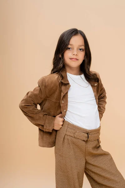 Brunette girl in brown suede jacket posing with hands on hips while looking at camera isolated on beige — Stock Photo