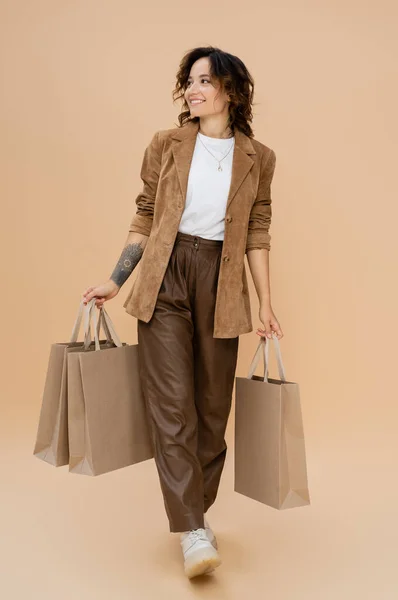 Donna gioiosa in giacca scamosciata che cammina con le borse della spesa e distoglie lo sguardo sullo sfondo beige — Foto stock