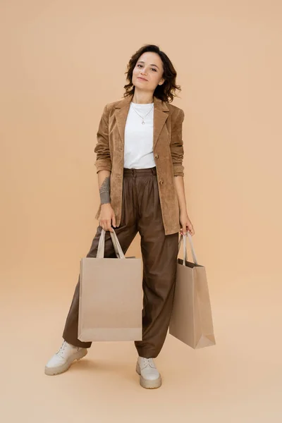 Pleine longueur de femme en tenue d'automne à la mode posant avec des sacs à provisions sur fond beige — Photo de stock