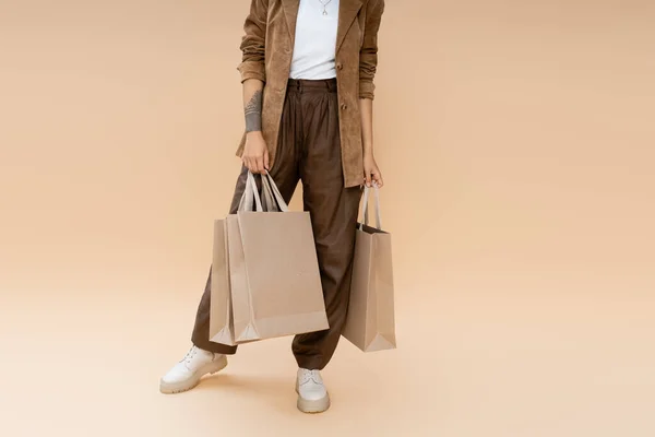 Visão parcial de mulher tatuada em calças marrom segurando sacos de compras no fundo bege — Fotografia de Stock
