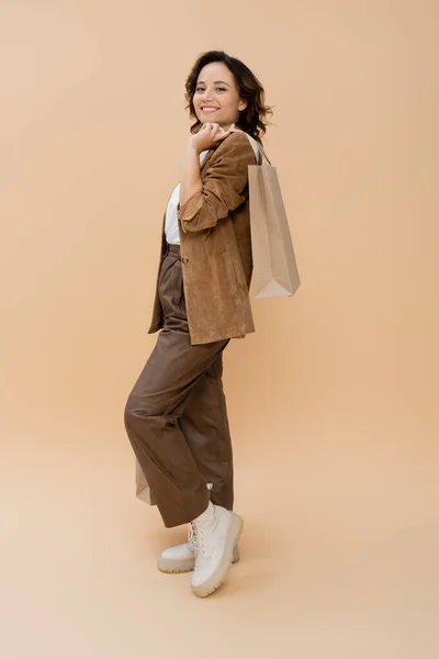 Полная длина улыбающейся женщины в коричневых брюках и замшевой куртке держа сумку с покупками на бежевом — стоковое фото