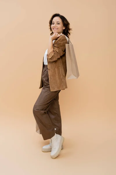 Full length of smiling woman in brown pants and suede jacket holding shopping bag on beige — Stock Photo