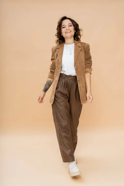 Full length of tattooed woman in suede jacket and brown pants walking on beige — Stock Photo