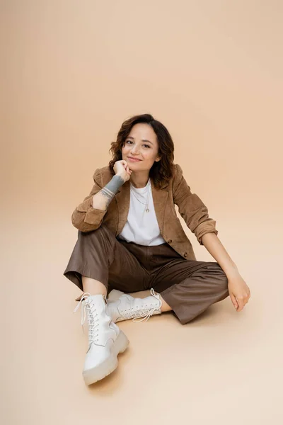 Morena mujer con el pelo ondulado con ropa de otoño de moda mientras se sienta con la mano cerca de la cara sobre fondo beige - foto de stock