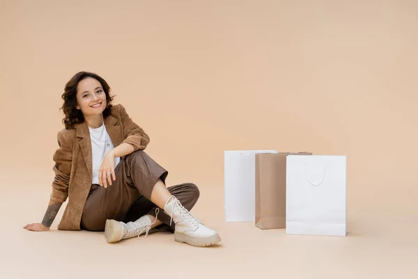 Щаслива жінка в стильному осінньому одязі та черевиках, що сидять біля сумки для покупок на бежевому фоні — стокове фото