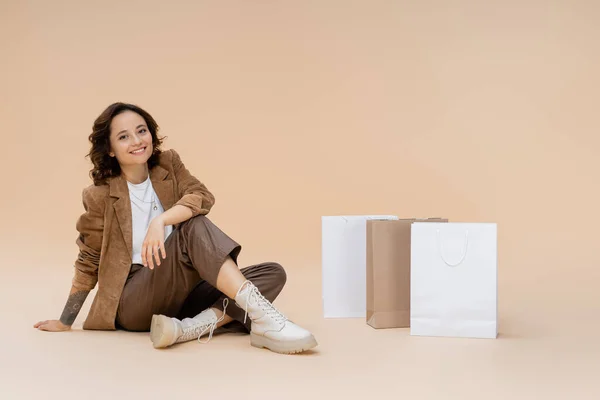 Mujer feliz en ropa de otoño elegante y botas sentadas cerca de bolsas de compras en fondo beige - foto de stock